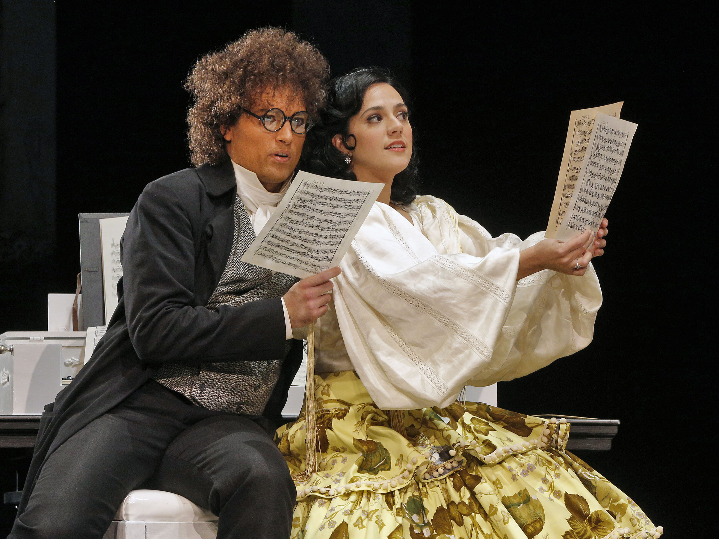A man with curly hair in a suit is sitting in a chair next to a woman wearing a white blouse and gold skirt. They are holding up paper scripts.