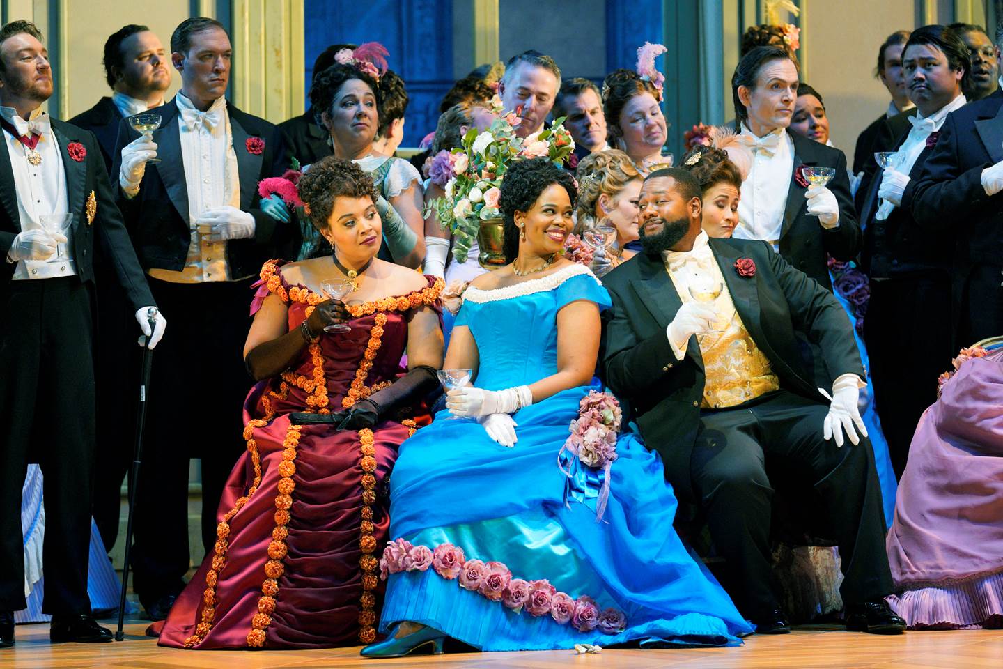 A group of opera performers in lavish 19th-century costumes on stage. Two women in elaborate gowns. sit at the forefront, engaging with each other. They are surrounded by elegantly dressed men and women in formal attire, all holding glasses and appearing to be in a celebratory mood.