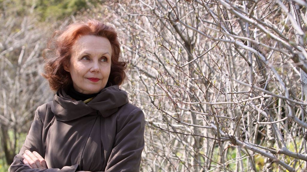 Kaija Saariaho in a brown top with her arms folded standing in front of bushes with no leaves.