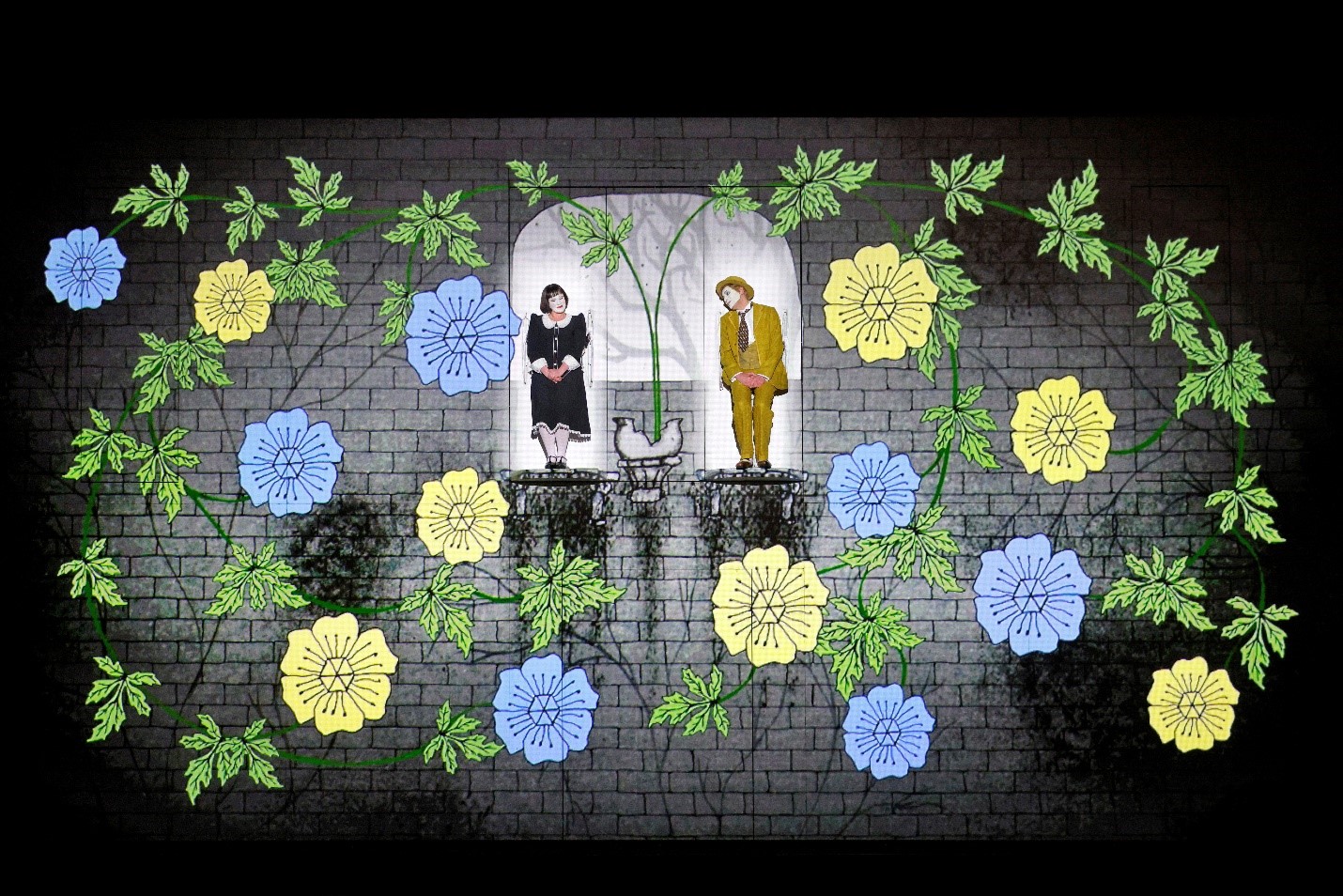 An artistic stage set featuring two performers standing in separate arched windows against a brick wall backdrop. The performer on the left is dressed in a black and white outfit, while the performer on the right is in a yellow suit with a matching hat. The scene is decorated with large, colorful flowers and green vines, with a whimsical and illustrated style. The performers appear to be interacting with each other through their body language, adding a playful and animated feel to the scene.