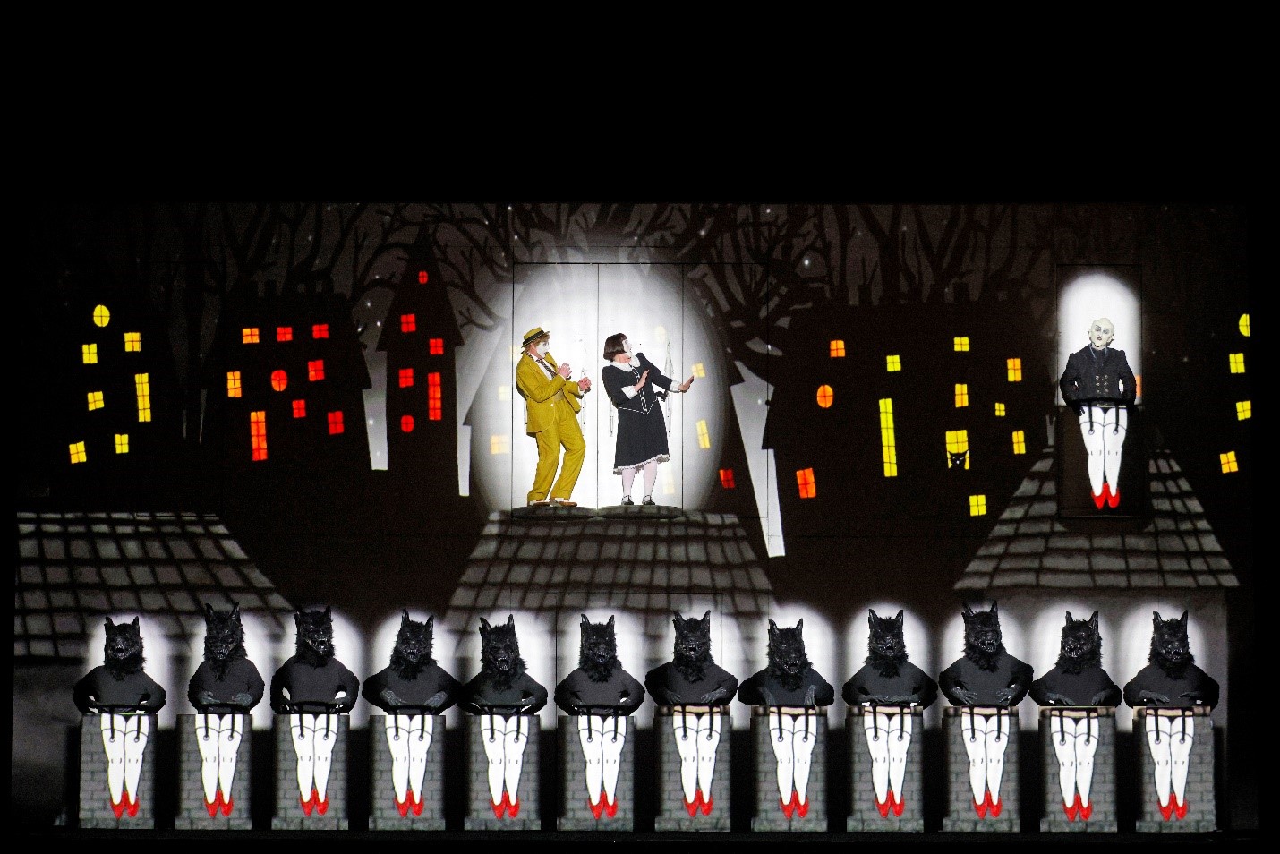 An elaborate stage scene featuring three performers and a chorus line. The central performers, dressed in a yellow suit and a black outfit, stand in separate windows with animated expressions. To the right, another performer dressed in black sits in a window. The foreground showcases a row of performers dressed as wolves with black tops, white leggings, and red shoes, standing atop pillars. The backdrop is a dark, whimsical cityscape with trees and buildings illuminated by colorful, glowing windows, creating a night-time ambiance.