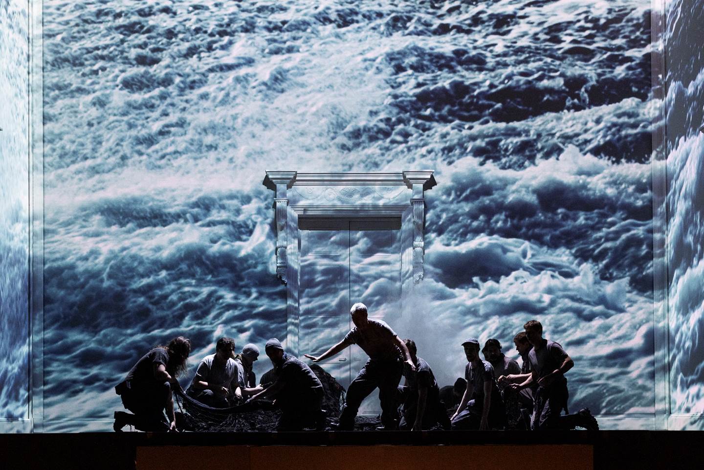 Silhouette&#39;s of several cast members on their knees with an image of ocean waves covering the wall behind them.