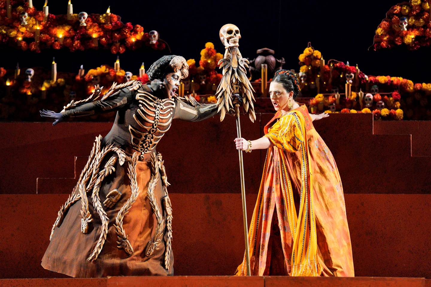 Scene from Frida of a woman in orange dress and a skeleton both holding onto a stick with a skull on top
