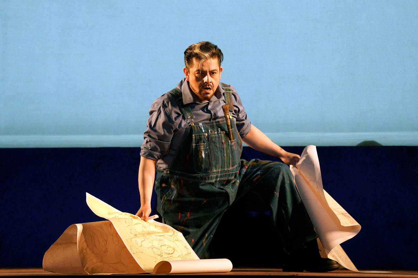 Scene from Frida A man in overalls sitting on a stage with a piece of paper.