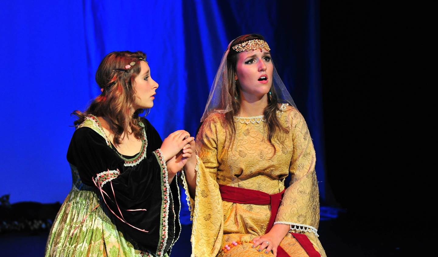 Two girls kneeling on stage. One has a distressed look on her face while the other one is holding her hand.