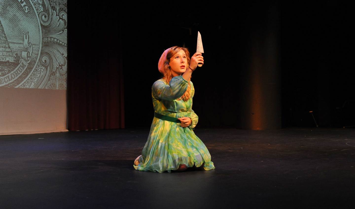 A girl in a green dress is on her knees holding a knife in the air as she looks at it with a concerned expression