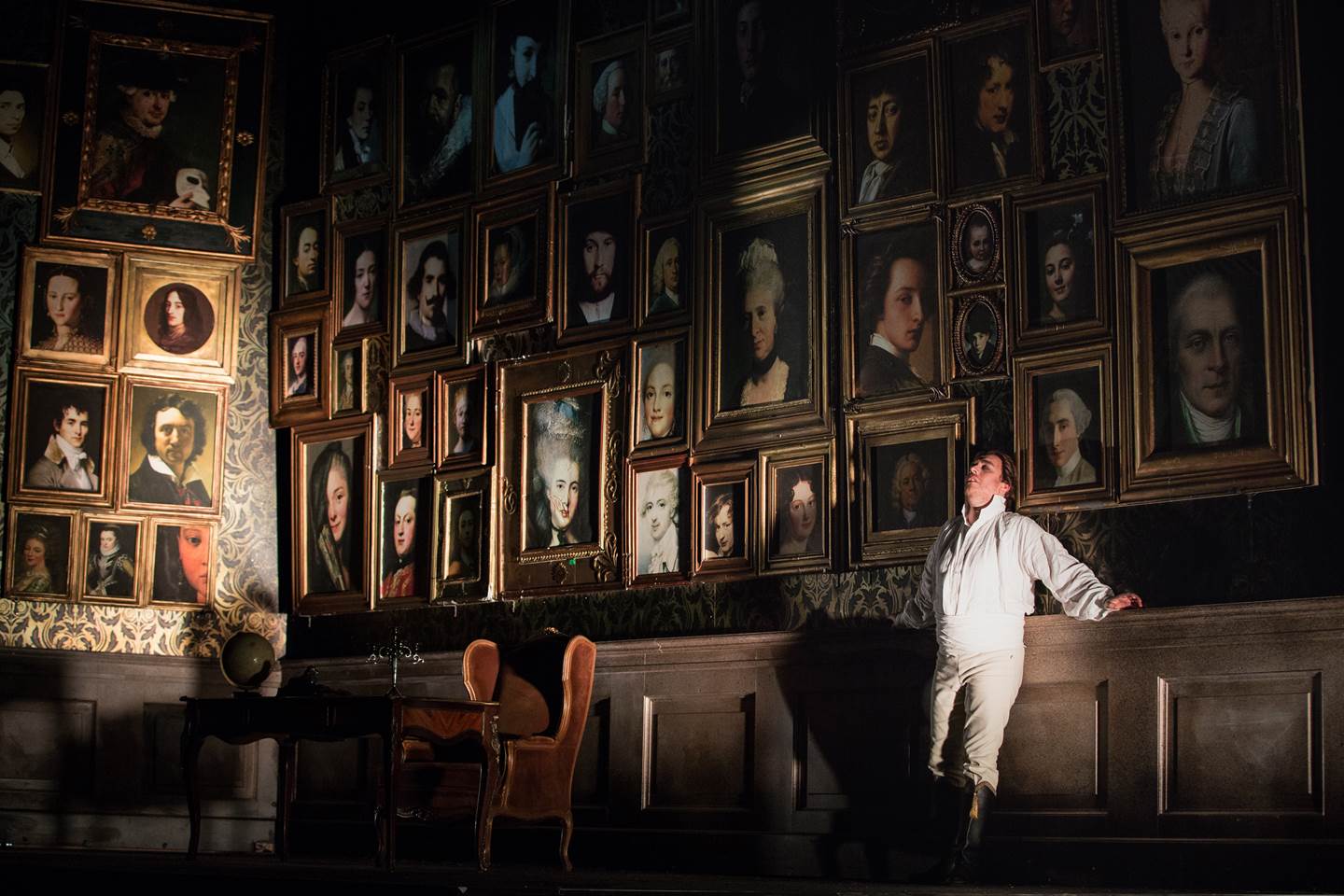 Un Ballo in Maschera male cast member standing against a large wall filled with framed portrait paintings.